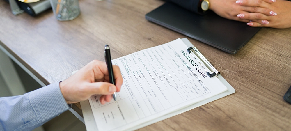 Client's hand filling out an insurance claim form on a clipboard during a meeting with an agent in an office setting